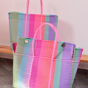 May include: Two rectangular tote bags constructed from woven plastic strips in a vibrant multicoloured striped pattern featuring shades of neon pink, light blue, lavender, and lime green, each equipped with two matching neon pink rope handles.
