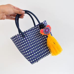May include: A small, woven tote bag in navy blue and white, featuring a decorative heart-shaped embellishment with a yellow tassel. The bag has navy blue handles and a square shape, perfect for carrying small items.
