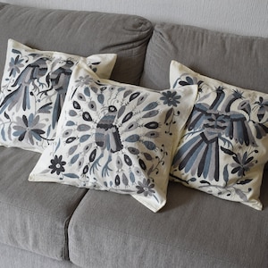 May include: Three decorative throw pillows with embroidered bird designs in shades of gray and blue on an off-white background. The pillows are square and rest on a gray sofa, adding a touch of elegance to the living space.