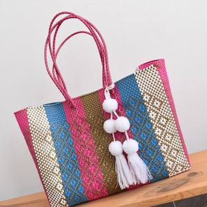 May include: A woven tote bag with pink, blue, and brown stripes and a white geometric pattern. The bag has pink handles and is adorned with white pom-poms and tassels. The bag is made of a durable, woven material.