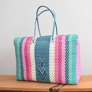May include: A rectangular tote bag constructed from woven plastic strips in a vibrant striped pattern. The design features vertical bands of teal, bright pink, light pink, and white. It includes two matching woven handles at the top.
