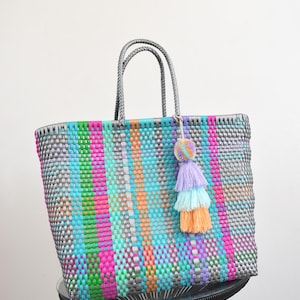 May include: A woven tote bag featuring a multi-coloured striped pattern in shades of silver, bright pink, lime green, turquoise, and orange. The bag has two braided silver handles and a decorative multi-layered tassel in lavender, light blue, and peach.