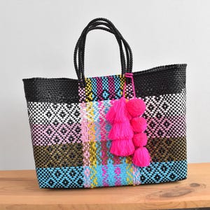 May include: A woven tote bag with a black top and handles, featuring a geometric pattern in white, pink, blue, and olive green. A cluster of bright pink pom-poms hangs from the handle. The bag sits on a wooden surface.