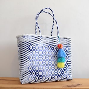 May include: A white and blue woven tote bag with a geometric diamond pattern. The bag has matching handles and a colourful tassel with red, blue, and yellow accents. The bag is sitting on a wooden surface.
