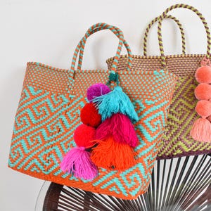 May include: Two woven tote bags with handles. One is orange with a turquoise geometric pattern and colourful tassel accents. The other is brown with a green and yellow geometric pattern and peach tassel accents. The bags are on a black and brown chair.