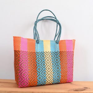 May include: A colorful woven tote bag with a rectangular shape. The bag features a vibrant mix of orange, pink, blue, and yellow stripes and diamond patterns. It has blue rope handles, perfect for carrying items. The bag is made of a durable, woven material.