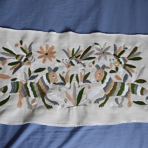 Puede incluir: Una funda de cojín de lino blanco con animales y flores bordados de colores. Los animales son estilizados y tienen un patrón a rayas. Las flores son en tonos de marrón, blanco y verde.