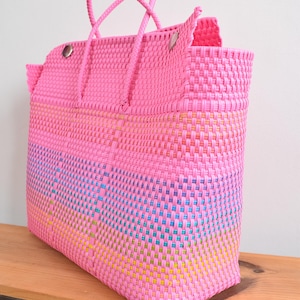 May include: A large tote bag constructed from woven pink plastic strips. The bag features two matching pink braided handles and silver-toned metal hardware at the top corners. The lower section incorporates a subtle gradient of blue, green, and yellow.