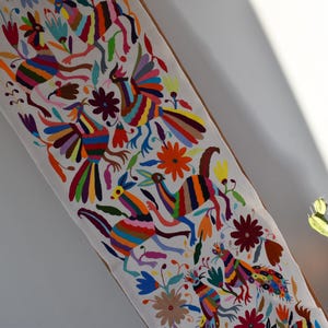 Puede incluir: Un camino de mesa de tela blanca con pájaros y flores bordados de colores vivos. Los pájaros se representan en una variedad de poses, incluyendo volando, posados y caminando. Las flores están en flor y tienen una variedad de colores, incluyendo rojo, amarillo, naranja y morado.