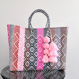 May include: A woven tote bag with a striped pattern in shades of pink, red, gray, and brown. The bag has a black and white striped handle and is adorned with pink pom-poms and a tassel. The bag is sitting on a glass table.