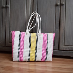 May include: A colorful tote bag with vertical stripes in pink, white, yellow, and black. The bag has white rope handles and is made of woven material. The bag is sitting on a wooden floor in front of a gray cabinet.