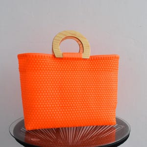 May include: A bright orange woven tote bag with a natural wood handle. The bag has a textured woven pattern.