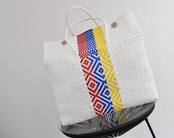 Könnte beinhalten: Weiße gewebte Tragetasche mit einem farbenfrohen vertikalen Streifen. Der Streifen weist rote, blaue und gelbe geometrische Muster auf. Die Tasche hat weiße Griffe und steht auf einem schwarzen Stuhl.
