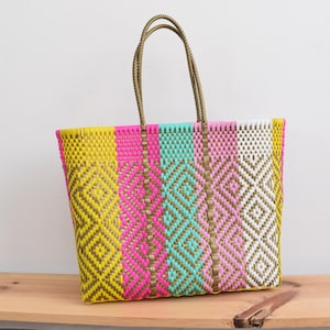 May include: A vibrant woven tote bag with a geometric diamond pattern. The bag features vertical stripes of yellow, pink, turquoise, and white. It has brown handles and is on a wooden surface.