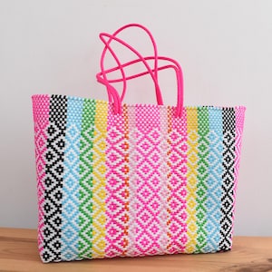 May include: A vibrant, rectangular tote bag with a woven design. The bag features a repeating diamond pattern in pink, black, white, blue, green, and yellow. It has bright pink handles. The bag is made of a durable, woven material.