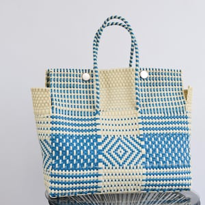 May include: A blue and cream woven tote bag with a geometric pattern. The bag has a diamond design on the front and two handles. It is secured with two white buttons. The bag is made of a durable, woven material, suitable for carrying items.