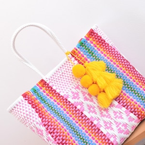 May include: A vibrant woven tote bag with a white handle and a yellow tassel. The bag showcases a pink and white diamond pattern with stripes of red, blue, and green. The bag is resting on a wooden surface.