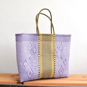 May include: A lavender woven tote bag with a central gold and olive green stripe. It has olive green handles. The bag is displayed on a wooden surface. The bag is approximately 30 cm wide.
