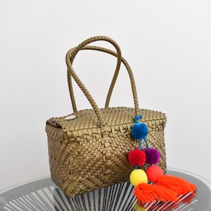 May include: A woven gold handbag with two handles and a colourful pom-pom tassel. The tassel is made of red, yellow, blue, and purple pom-poms.