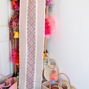 Puede incluir: Un camino de mesa tejido de color rosa y blanco con flecos, colgado de una escalera de madera con pompones de colores y bolsas tejidas.