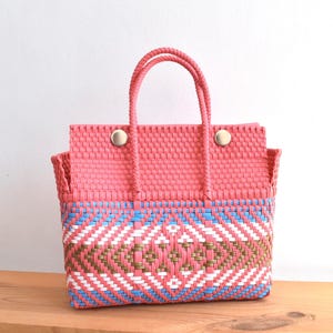May include: A woven tote bag in coral pink with a geometric pattern in blue, white, and gold. The bag has two handles and is secured with two gold-colored buttons. The bag is sitting on a wooden surface.