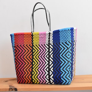 May include: A brightly coloured woven tote bag with a geometric design. The bag displays a vibrant mix of colours including red, orange, yellow, pink, white, blue, and black. It has two black and white striped handles.