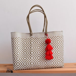 May include: A woven tote bag with a geometric diamond pattern in white and olive green. The bag has olive green handles and a red tassel. The bag is sitting on a wooden surface.