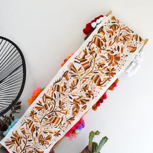 Puede incluir: Textil bordado de color crema con un diseño floral y animal en tonos marrón, naranja y amarillo. Los bordes están adornados con pompones de colores. Un elemento decorativo para el hogar, de unos 150 cm de largo.