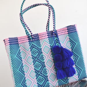 May include: A woven tote bag with pink, blue, and green geometric patterns. The bag has blue pom-poms attached to the handles.