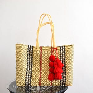 May include: Woven tote bag with a geometric pattern in beige, black, and red. The bag has beige rope handles and a red tassel. The bag is designed for carrying items.