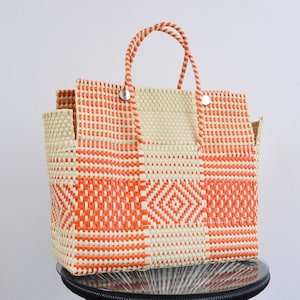 May include: A woven tote bag featuring a geometric pattern in bright orange and cream. The bag has two matching woven handles and a rectangular base structure with a repeating diamond motif integrated into the centre of the front panel design.
