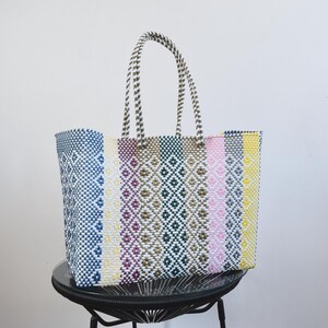 May include: A rectangular tote bag constructed from woven plastic strips featuring a multi-coloured vertical striped pattern in shades of blue, purple, green, pink, and yellow, accented with white diamond motifs and two matching woven handles.
