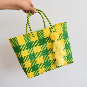 May include: A woven tote bag featuring a bright yellow and green houndstooth pattern. The bag has two braided handles in matching yellow and green colours and is decorated with a large, fluffy yellow tassel attached to one of the handles.