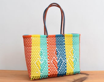 Könnte beinhalten: Eine farbenfrohe, rechteckige Tragetasche aus gewebtem Material. Sie hat vertikale Streifen in Orange, Gelb, Türkis und Marineblau. Die Griffe sind eine Kombination aus Orange und Marineblau. Die Tasche besteht aus strapazierfähigem, gewebtem Material.
