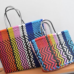 May include: Two woven tote bags in bright colours. The bags have a zig-zag pattern in shades of red, orange, yellow, pink, white, black, and blue. The handles are black and white. The bags are made of a durable, woven material.