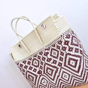 May include: Woven tote bag with a geometric pattern in burgundy and cream. The bag has a rectangular shape with a cream-coloured top and handles. The bag is made of a woven material and is suitable for carrying items.