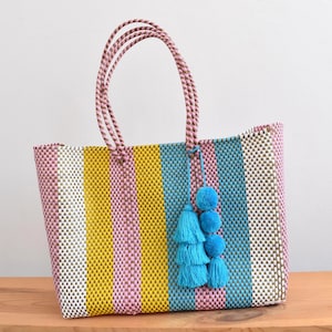 May include: A colorful tote bag with vertical stripes in yellow, pink, white, and blue. The bag has pink rope handles and a blue tassel with pom-poms. The bag is made of woven material.