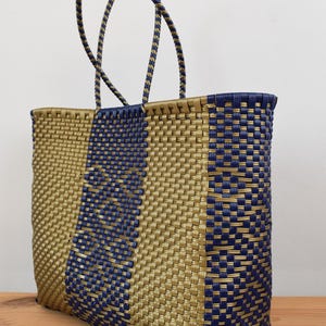 May include: Gold and navy blue woven tote bag. The bag has a rectangular shape with a woven pattern. Twisted rope handles in matching colours are attached. The bag is designed for carrying items.
