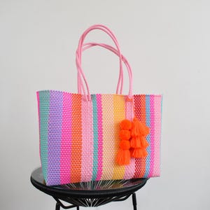 May include: A rectangular tote bag woven from plastic strips in a vibrant vertical striped pattern of pink, orange, yellow, purple, and blue. The bag features two pink braided handles and a decorative charm consisting of three orange pom-poms and tassels.