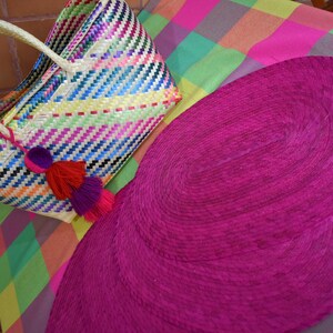 Op de afbeelding: Twee ovale roze geweven placemats op een kleurrijk geruit tafelkleed. Een grote, kleurrijke geweven draagtas met een witte handvat en kwastjes ligt op het tafelkleed.