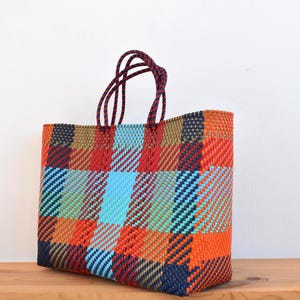 May include: A colorful woven tote bag with a plaid pattern in shades of orange, red, blue, and brown. The bag has a rectangular shape and dark red handles. The bag is sitting on a wooden surface.