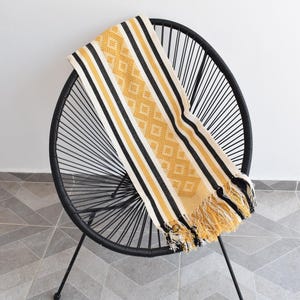 Puede incluir: Una silla de alambre negra con una manta tejida con estampado amarillo, negro y blanco sobre el asiento. La manta tiene un patrón de diamantes y flecos.