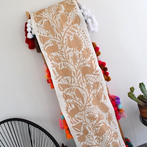 Puede incluir: Un largo camino de mesa de tela blanquecina con un patrón repetido de animales, flores y follaje de color marrón. El camino de mesa está drapeado sobre un marco de madera decorado con pompones y borlas de colores. Una silla negra y un cactus en maceta están en el fondo.
