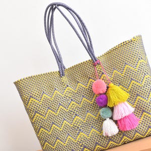 May include: A woven tote bag with a yellow and gray zigzag pattern. The bag has gray braided handles and is adorned with a colorful tassel of pink, yellow, purple, green, and white pom-poms. The bag is sitting on a wooden surface.