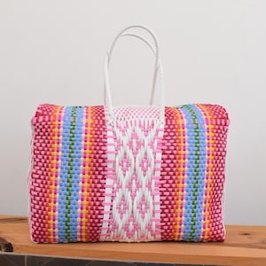 May include: A rectangular woven tote bag featuring a vibrant multi-coloured pattern of pink, red, yellow, blue, and green plastic strips. The bag includes a central white panel with a diamond-shaped weave design and two white braided handles.
