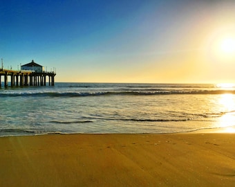May include: A pier extends out into the ocean with a golden sunset in the background. The water is a light blue and the sand is a light brown.