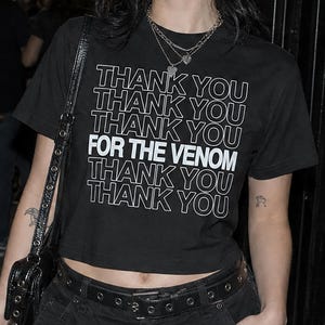 Puede incluir: Una camiseta negra de manga corta con el texto blanco THANK YOU repetido cinco veces y la frase FOR THE VENOM centrada en letras blancas en negrita. La camiseta tiene cuello redondo y dobladillo corto, combinada con pantalones cargo negros.