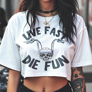 Op de afbeelding: Een wit gecropped t-shirt met een zwarte schedelprint en de tekst "Live Fast Die Fun". Het shirt heeft een motorstuurontwerp met de tekst "Live Fast" boven de schedel en "Die Fun" onder de schedel.
