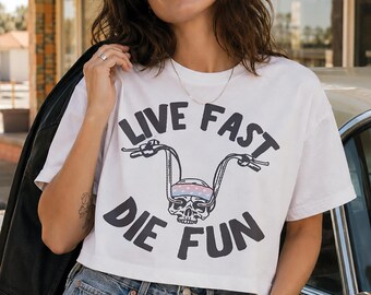 May include: A white short-sleeved t-shirt featuring a black graphic print of a skull wearing a patterned bandana with motorcycle handlebars extending from the eye sockets, accompanied by the black text "LIVE FAST" above and "DIE FUN" below.