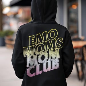 Op de afbeelding: Zwarte hoodie met de tekst "EMO MOMS MOM CLUB" in een verloop van geel, roze en wit. De hoodie wordt gedragen door een persoon die de rug naar de camera toekeert.